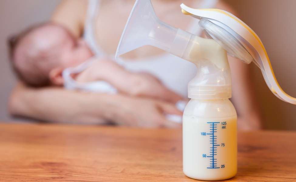 Seorang ibu sedang menggendong bayinya dan ada power pumping dan botol susu yang terisi penuh.